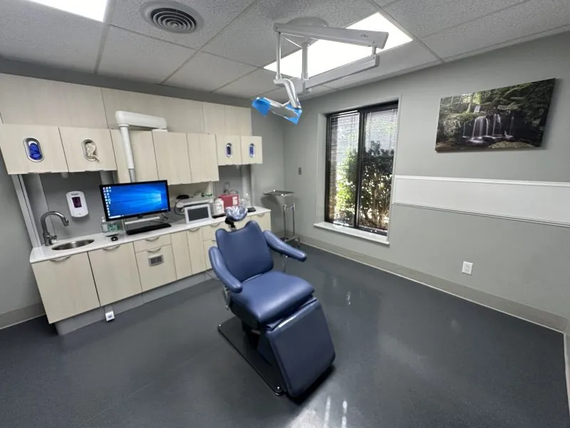 Dental treatment room with grey flooring, blue patient chair, white cabinetry, computer workstation, window with natural light, and wall-mounted monitor.