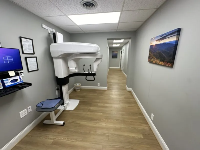 Dental treatment room with grey flooring, blue patient chair, white cabinetry, computer workstation, window with natural light, and wall-mounted monitor.