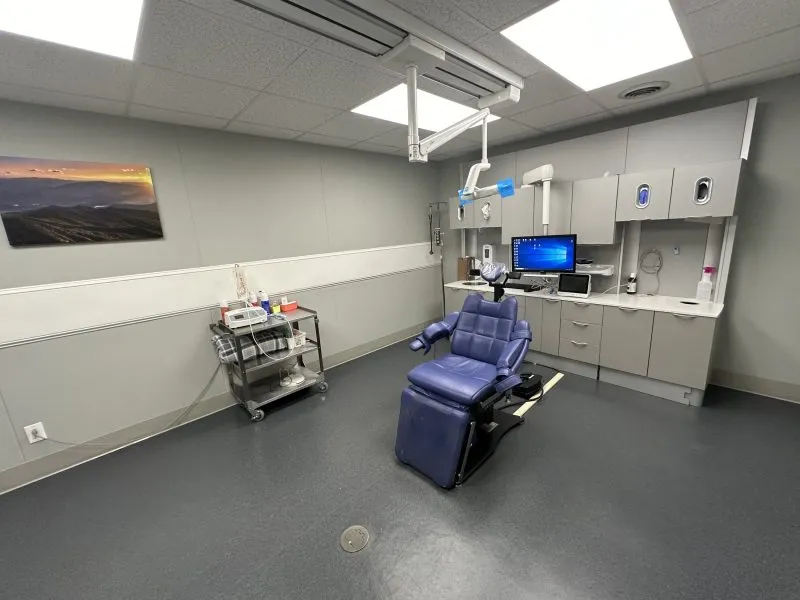 Modern dental treatment room equipped with blue patient chair, overhead dental delivery system, white cabinetry, grey flooring, and artwork.